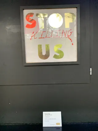 a framed placard that reads 'stop killing us' on a black wall