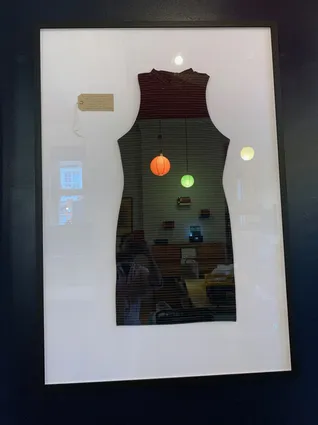 a framed black sleeveless dress on a white background with a brown swing tag next to it