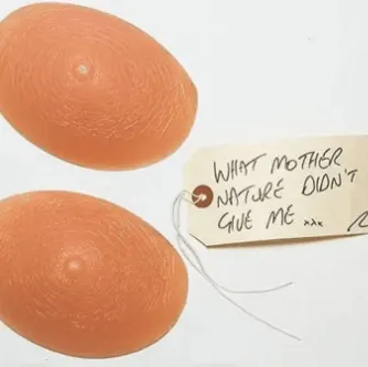 A pair of prosthetic breasts with a brown tag that reads 'what mother nature didn't give me xxx'
